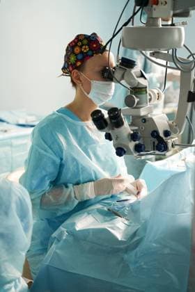 Adult female doctor in sterile mask and ornamental medical cap looking through surgical microscope against crop coworker in hospital, Model released SergeyMironov_Surgery_41.jpg Copyright: xSergeyxMironovx MR