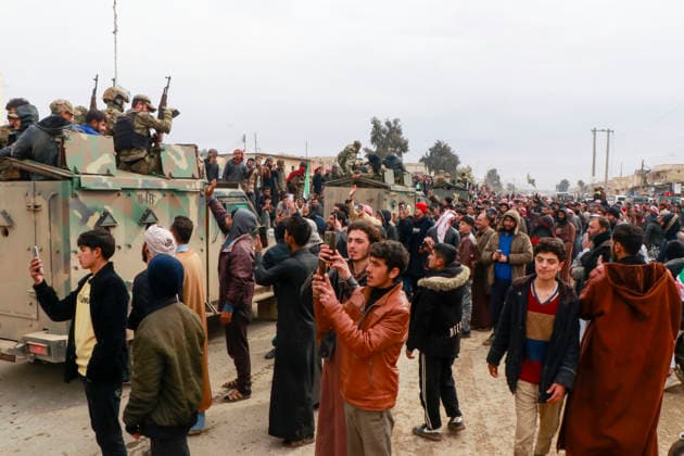 **Östliches Umland von Aleppo, Gouvernement Rakka, Syrien, Asien.** Große Menschenmenge versammelt sich entlang einer Straße, während gepanzerte Militärfahrzeuge mit bewaffneten Kämpfern in Tarnuniform langsam passieren. Zahlreiche Zivilisten filmen und fotografieren die Szene mit Mobiltelefonen, einige heben die Hände oder gestikulieren in Richtung der Fahrzeuge. Dichte urbane Umgebung mit Wohngebäuden, Strommasten und Bäumen im Hintergrund. Handlung: Ankunft und Durchfahrt militärischer Einheiten, Reaktionen der Bevölkerung. Perspektive: Weitwinkel, leicht erhöht, seitlich-frontal. Schlagworte: News, Politik, Sicherheit, Militär, Konflikt, Gesellschaft. Copyright: xAbdalhayxAbdalhayx