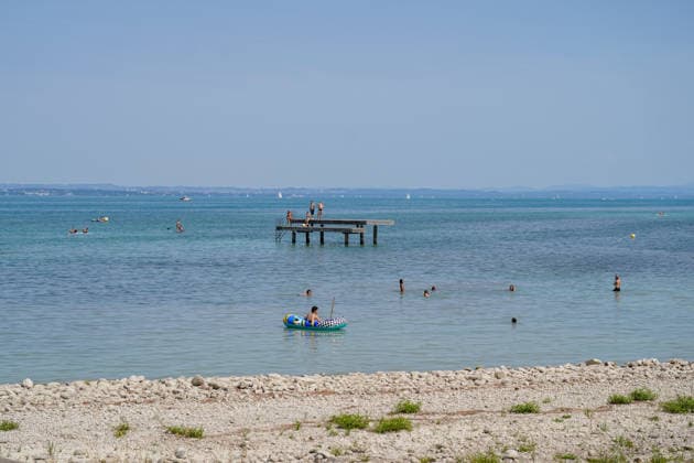 Bodensee: Badegäste und Wassersportler genießen einen sonnigen Sommertag im Bodensee. Menschen schwimmen im See, fahren mit Schlauchboot und Stand-up-Paddle; weitere Personen stehen auf einem Badesteg. Im Hintergrund Segelboote und das gegenüberliegende Ufer. Sommer, Hitze, Freizeit, Baden, Schwimmen, Wassersport, Tourismus, Ferien, Erholung, See, Strand, Badesteg, SUP, Panorama, Totale, Symbolbild. Copyright: xJOERANxSTEINSIEKx