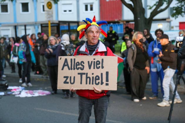 Berlin, Deutschland 24.09.2026: Teilnehmer der Protestkundgebung gegen die Verleihung des Axel Springer Awards an Peter Thiel hält ein Schild mit der Aufschrift Alles viel zu Thiel! und trägt eine bunte Narrenkappe. Im Hintergrund Demonstranten und eine Palästina-Fahne. Demonstration, Protest, Peter Thiel, Palantir, Axel Springer Award, Protestplakat, Wortspiel, Palästina, Aktivismus, Kundgebung, Porträt. Copyright: xAlonxDavidx