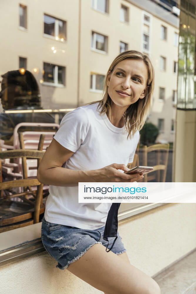 Portrait of woman with smartphone and protective mask in her hand waiting in front of a coffee shop model released Symbolfoto MFF05638