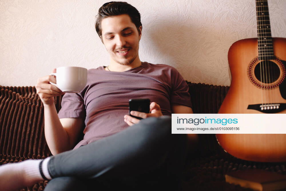 Happy young man using smart phone while drinking tea at home CR_DMBE200812-379179-01 ,model released, Symbolfoto