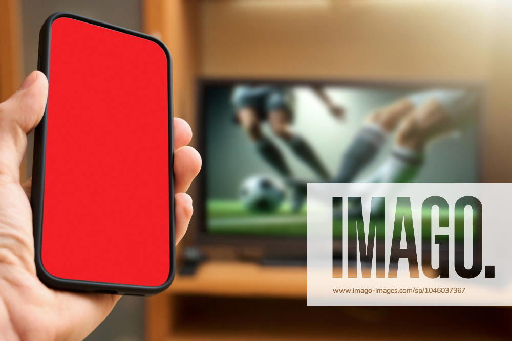 Hand holding a smartphone with a red screen in front of a TV showing a soccer match. Symbolic image of a red card in soccer. PHOTOMONTAGE