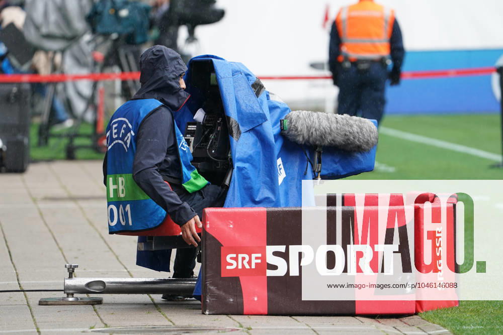 Kameramann bei einer UEFA-Veranstaltung, SRF SRG SSR, Regenwetter, Kamera mit Wetterschutz, Sportübertragung, Live-Berichterstattung, Fußballspiel, Medienberichterstattung, Fernsehproduktion, Kameratechnik, Sportjournalismus, Sicherheitskräfte im Hintergrund, Sportevent, TV-Sender, Broadcasting, Schweiz, Stadion, Außenübertragung, UEFA HB (Host Broadcaster), Sportreporter - Symbolbild