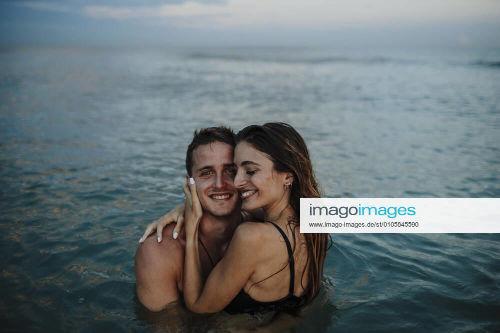 Smiling couple embracing while standing in water at beach during sunset model released Symbolfoto GMLF00721