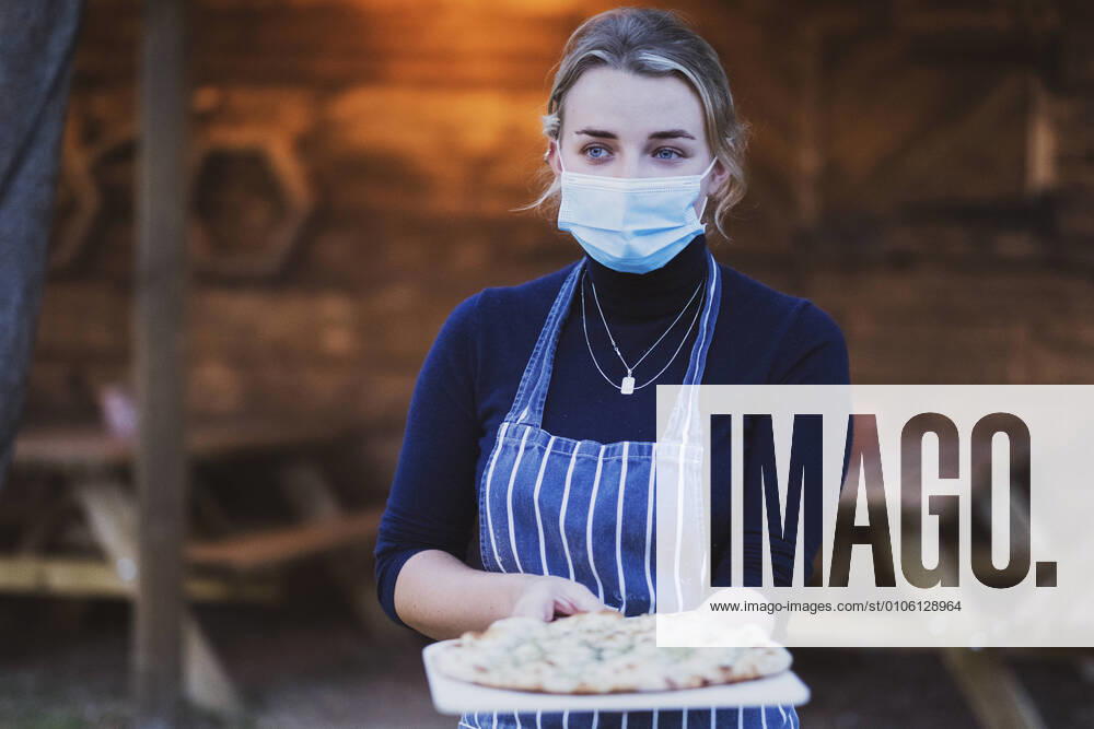 IMAGO Creative: Landleben - Essen Woman waitress in apron and face mask holding fresh pizza on a board Pizza Restaurant,United Kingdom,Woman waitress in apron and face mask holding fresh pizza on a board Woman waitress in apron and face mask holding fresh pizza on a board