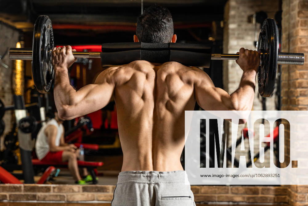 Rückenansicht eines jungen Bodybuilders, der seine Muskeln mit einer Langhantel vor einem Spiegel im Fitnessstudio anspannt Rückenansicht eines jungen Bodybuilders, der seine Muskeln mit einer Langhantel vor einem Spiegel im Fitnessstudio anspannt (License=RF) 23435157 ,model released, Symbolfoto