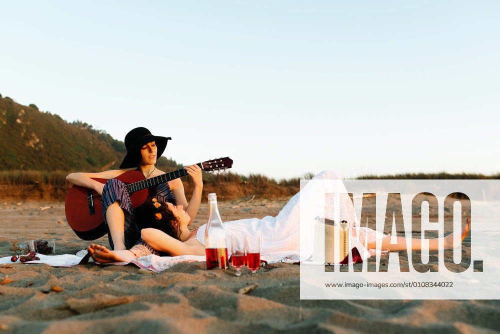 IMAGO Creative: Pride Month, Juni 2022, P?rchen und Familien Talented female musician playing acoustic guitar for girlfriend during picnic at seaside at sunset, Model released