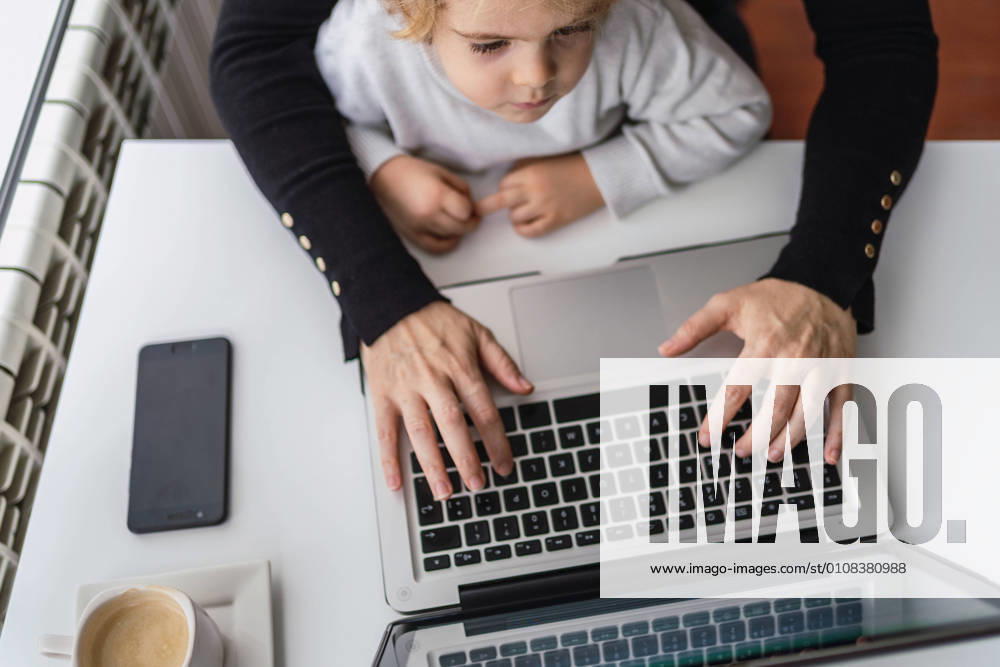 IMAGO Creative: Kreative Renaissance -- der junge Entrepreneur From above of crop anonymous female remote employee holding curious little child on knees while sitting at table and working with laptop at home