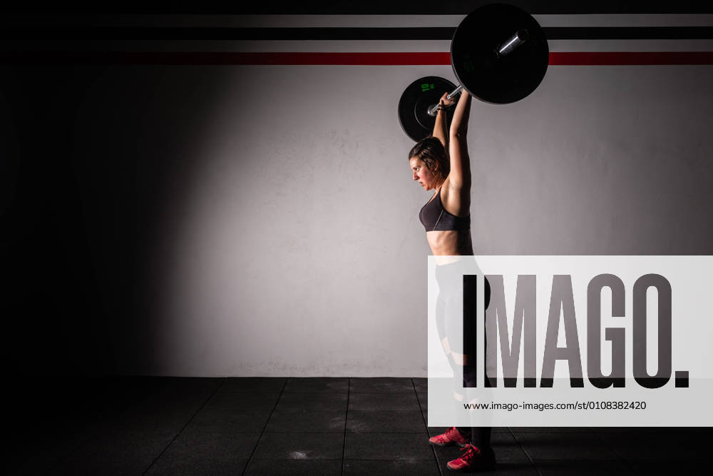 IMAGO Creative: 2021 Favoriten Side view of athletic young concentrated lady in sportswear upping barbell above head in gym
