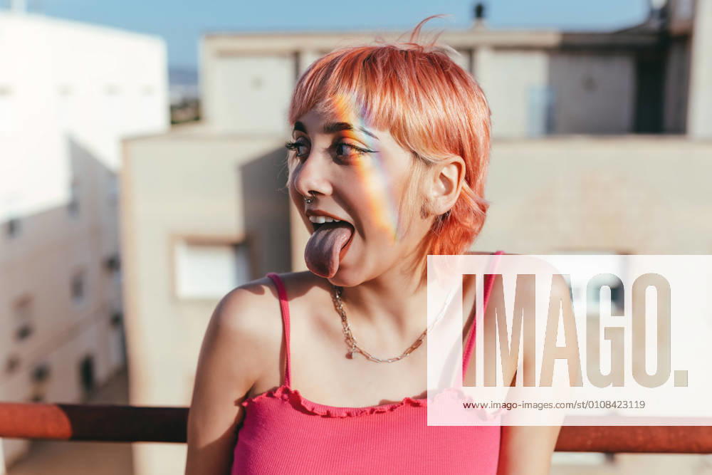 IMAGO Creative, Lifestyle, Barbiecore Cheerful female in summer wear looking away while sticking tongue out and leaning on railing in city