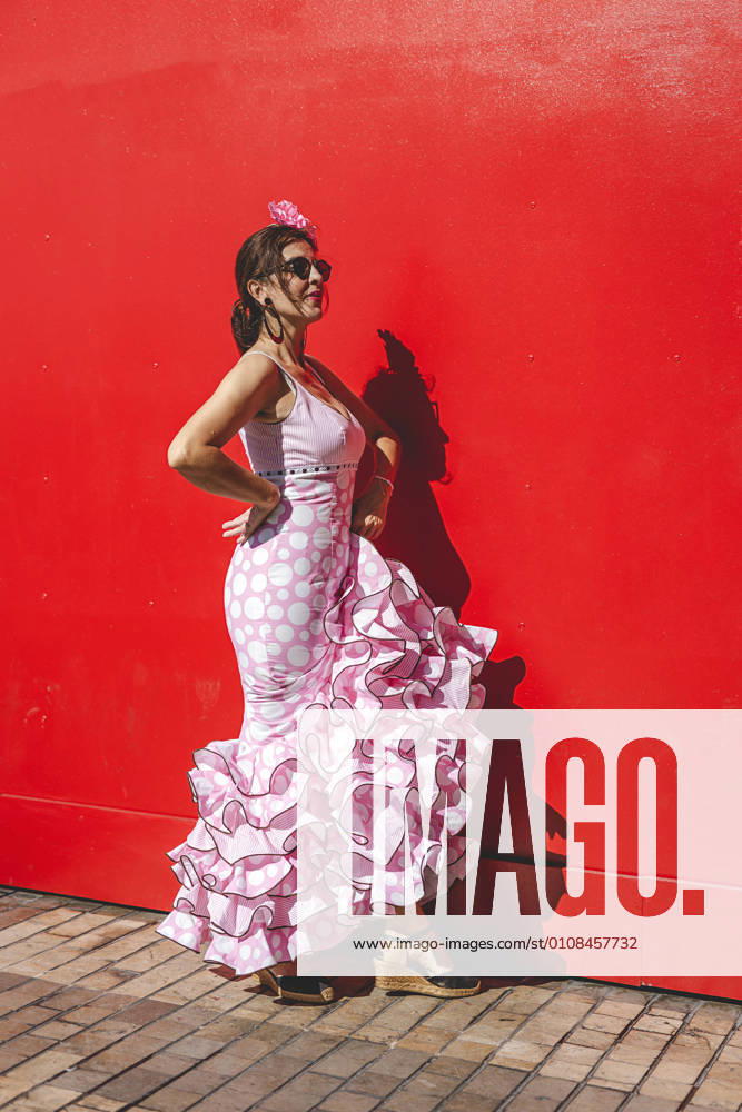 Side view of eccentric cheerful woman in colorful pink costume smiling and dancing by red wall background on sunny day
