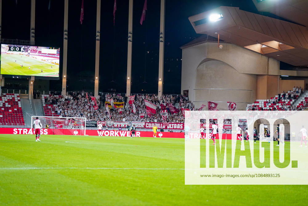 Fussball UEFA Conference League 2026 - 2027, 2. Spieltag - AS Monaco vs SC Freiburg - Stadion Louis II (Monaco): Blick vom Spielfeld auf den Gästeblock mit zahlreichen Fans des SC Freiburg, die ihre Mannschaft mit rot-weissen Fahnen, Fanbannern und Transparenten unterstützen. Im Vordergrund Spieler beider Mannschaften während einer Spielszene, im Hintergrund eine grosse Stadionanzeigetafel mit dem Spielstand 0:0. Auswärtsfans, Fankultur, Ultras, Fanblock, Zuschauer, Tribüne, Stimmung, Anfeuerung, Fahnenmeer, Hintertorperspektive, Europapokal, Stadion, Totale.