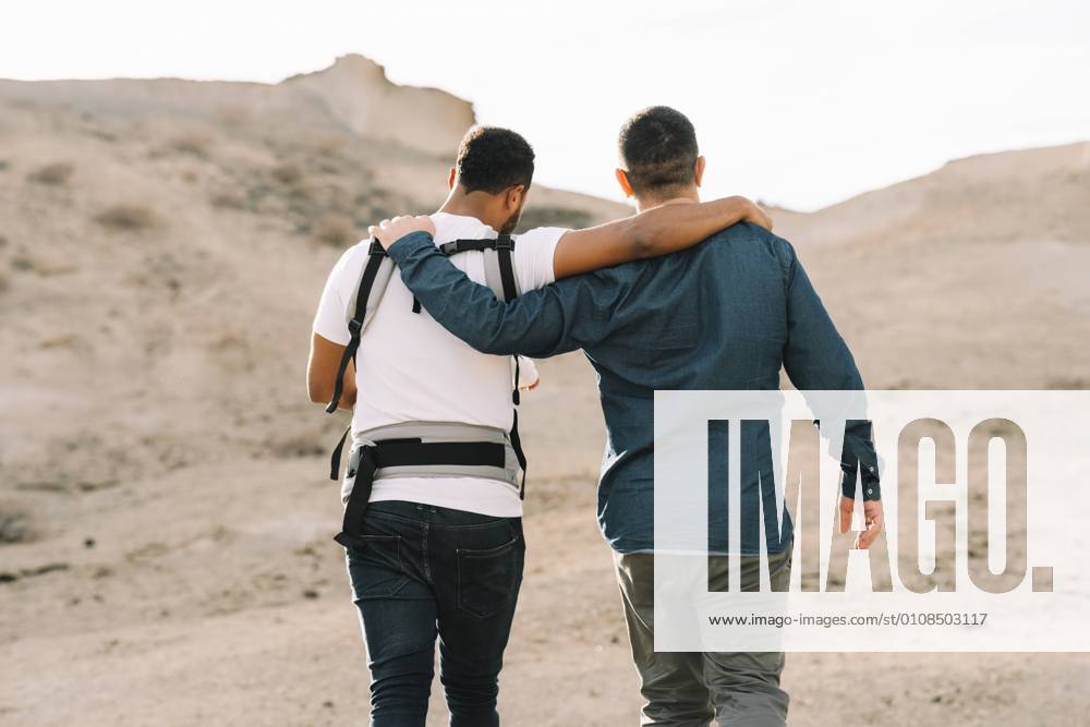 IMAGO Creative: Pride Month 2022, Engagement der Jugendlichen Back view of cropped black casual man holding on shoulder anonymous male boyfriend and holding little calm baby in grey carrier while strolling on nature at daytime, Model released