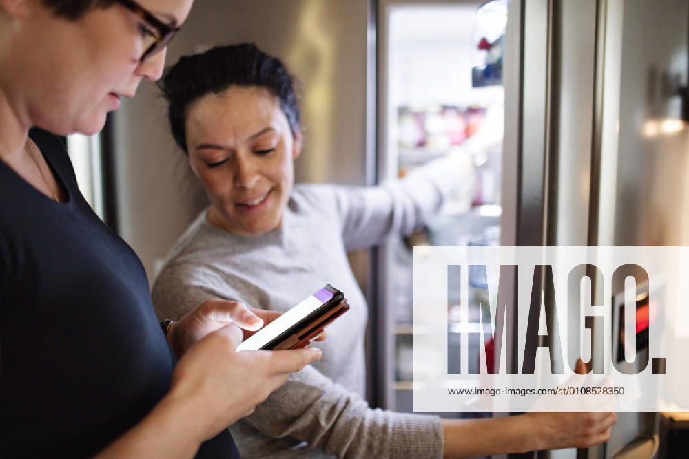 Woman showing mobile phone while girlfriend opening refrigerator in kitchen , model released, property released