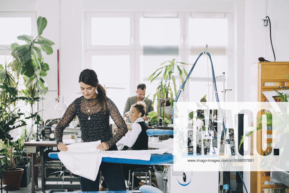 IMAGO Creative: Kreative Renaissance -- der junge Entrepreneur Female entrepreneur ironing white fabric while colleagues working in background at creative office , model released, property released