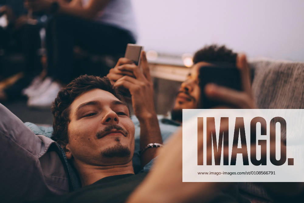 IMAGO Kreativ: Urbane Kultur - Sommer auf den Dächern Young male friends using smart phones while lying on terrace at dusk , model released, property released
