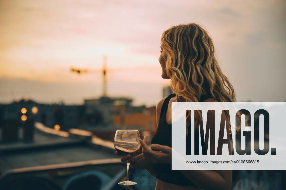 IMAGO Kreativ: Urbane Kultur - Sommer auf den Dächern Thoughtful young woman having wine while looking away on terrace during rooftop party at sunset , model released, property released