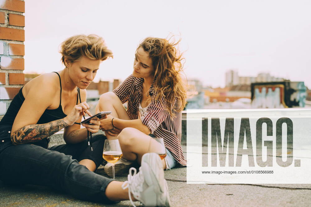 IMAGO Creative: Jahreszeit Frühling, Picknick Woman showing mobile phone to friend while sitting on terrace during rooftop party , model released, property released