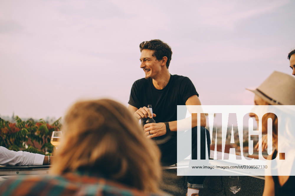 IMAGO Kreativ: Urbane Kultur - Sommer auf den Dächern Smiling man looking away while enjoying beer with friends at rooftop party against sky , model released, property released