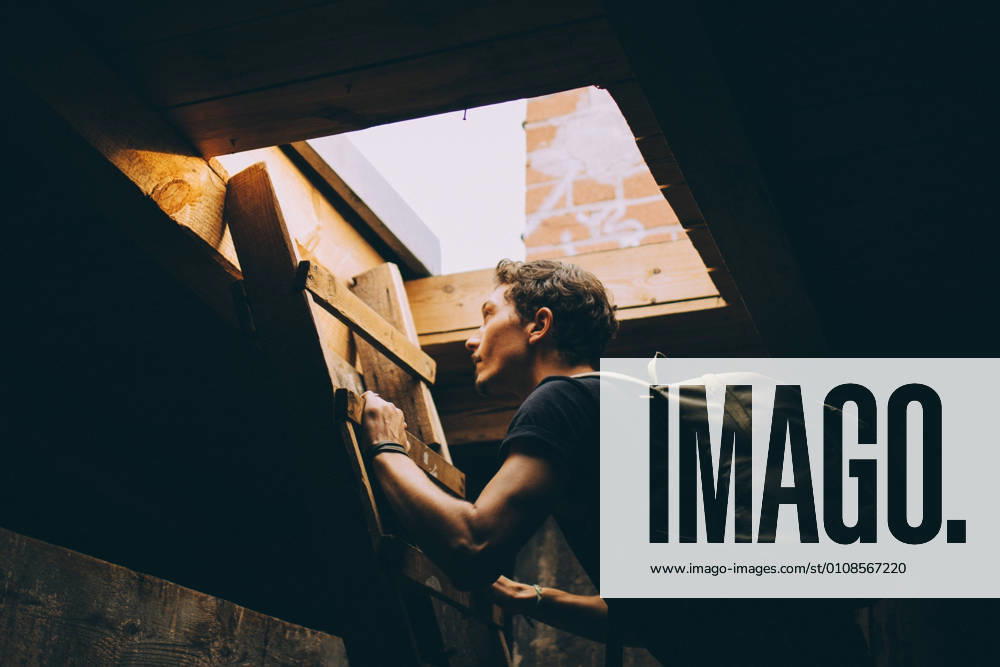IMAGO Kreativ: Urbane Kultur - Sommer auf den Dächern Side view of young man carrying backpack while climbing on ladder towards terrace , model released, property released
