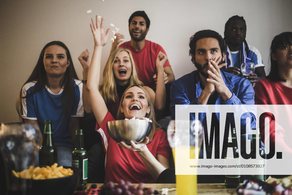 IMAGO Creative, Lifestyle, Leben in einer Wohngemeinschaft, WG Male and female soccer fans watching match while sitting on sofa at home , model released, property released