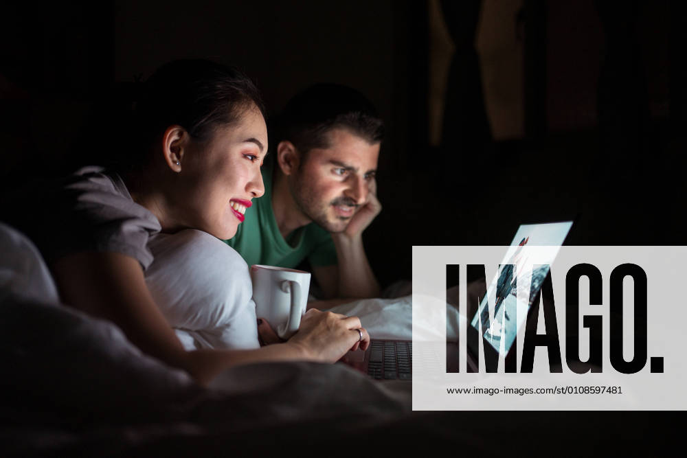 Smiling Asian female with cup of hot drink lying on bed near content boyfriend in casual wear while watching film on netbook at home