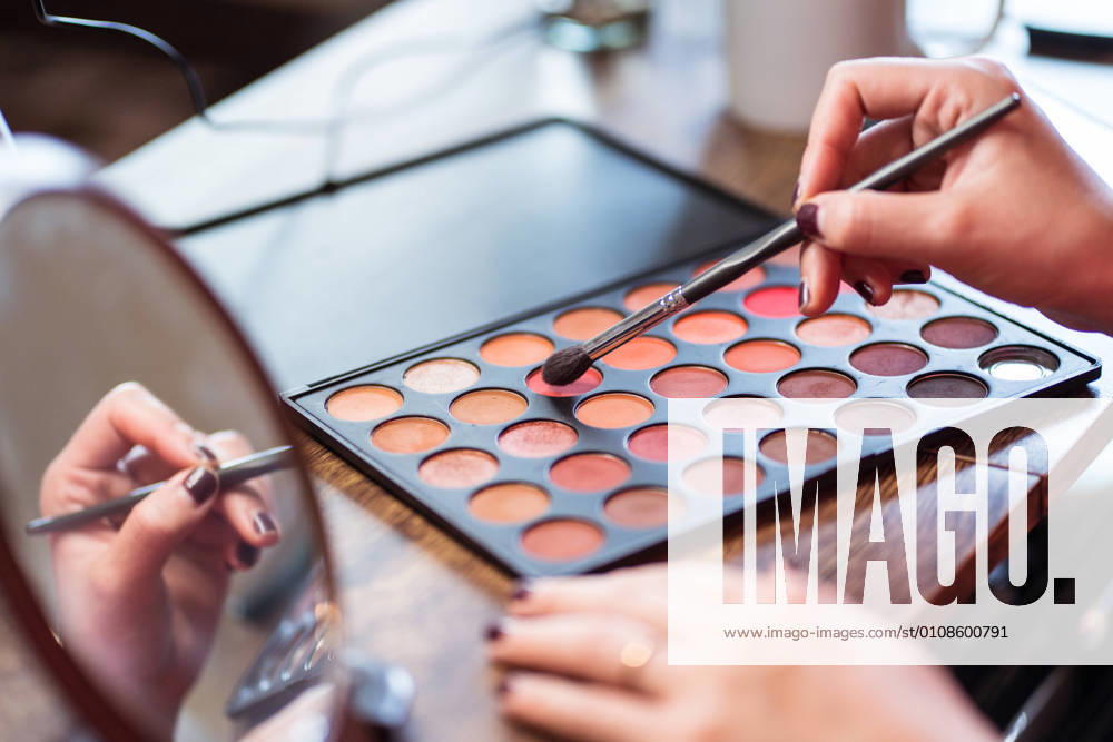 Crop anonymous female beauty blogger with colorful palette applying makeup while sitting at table in front of mirror and recording video tutorial