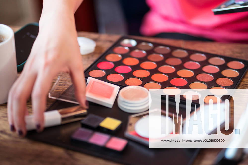 Crop anonymous female beauty blogger with colorful palette applying makeup while sitting at table in front of mirror and recording video tutorial
