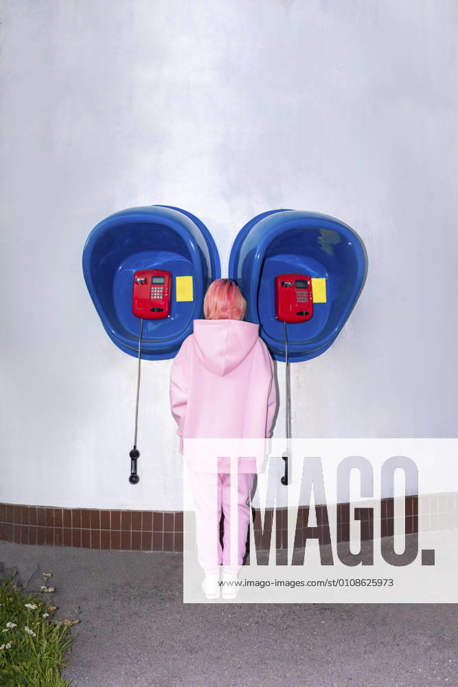 IMAGO Creative, Lifestyle, Barbiecore Rear view of young woman with pink hair wearing pink hooded shirt standing in front of telephone booths model released Symbolfoto VPIF03277