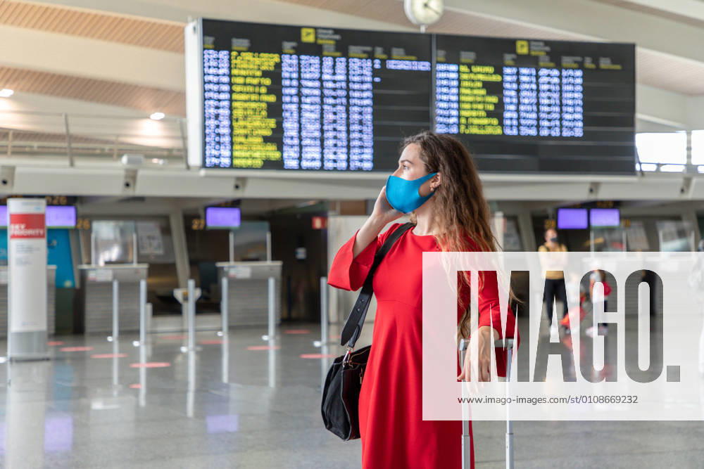 IMAGO Creative: Welttourismustag am 27. September Serious female traveler in mask standing with suitcase on background of departure board and talking on the phone while waiting for flight