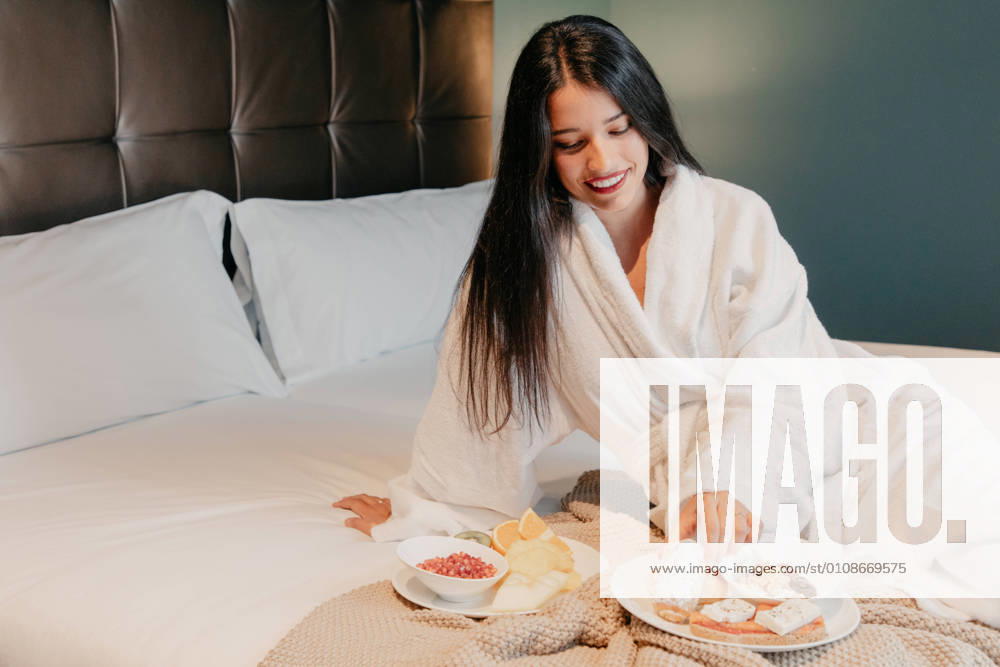 Happy relaxed young female in white bathrobe sitting on bed and enjoying delicious breakfast in hotel room