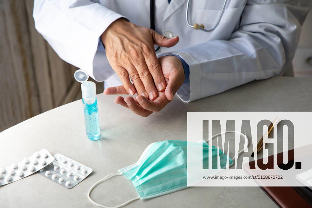 Male therapist in medical gown sitting at table in hospital and sanitizing hands with antiseptic while preparing for treatment of patients during coronavirus outbreak
