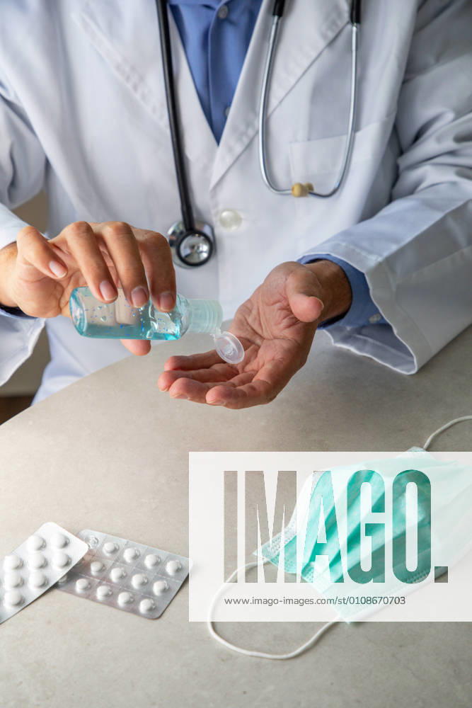 Male therapist in medical gown sitting at table in hospital and sanitizing hands with antiseptic while preparing for treatment of patients during coronavirus outbreak