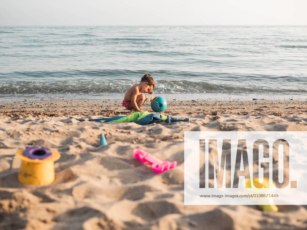 Side view of cute little girl with pigtails trying to make sand castle while playing on beach near waving sea on resort