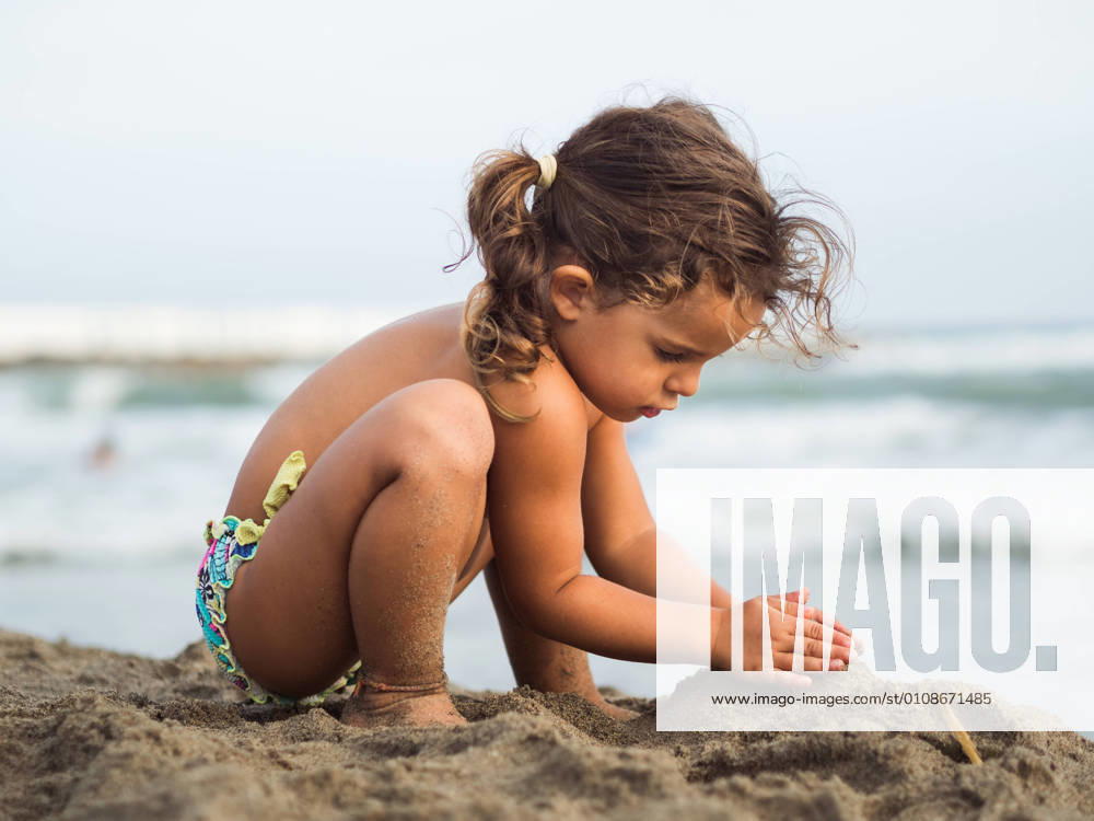 Side view of cute little girl with pigtails trying to make sand castle while playing on beach near waving sea on resort