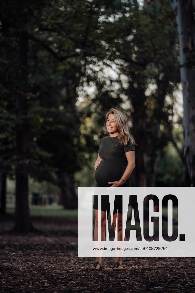 Happy pregnant woman in casual dress stroking belly while standing on pathway in park with green trees in sunny summer day with blue sky, Model released