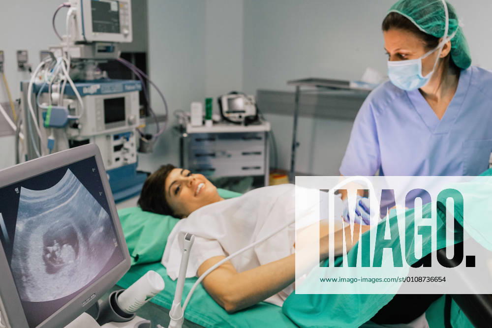 Female doctor in sterile mask and blue glove using ultrasound scanner while examining cheerful pregnant woman and looking at computer screen in hospital, Model released, Property released