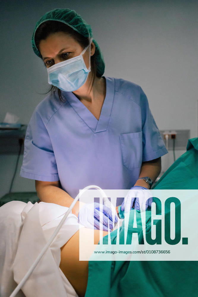 Female doctor in sterile mask and blue glove using ultrasound scanner while examining pregnant woman and looking at computer screen in hospital, Model released, Property released