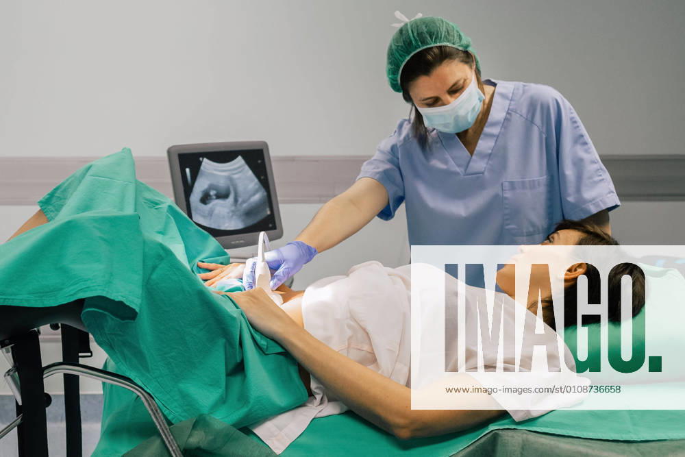 Female doctor in sterile mask and blue glove using ultrasound scanner while examining cheerful pregnant woman in hospital, Model released, Property released