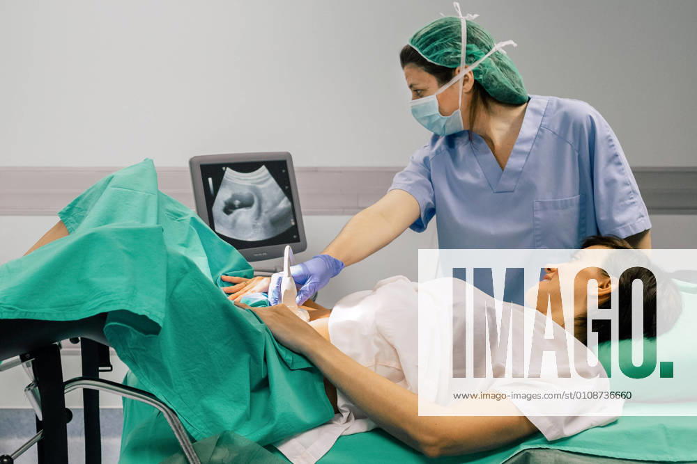 Female doctor in sterile mask and blue glove using ultrasound scanner while examining cheerful pregnant woman and looking at computer screen in hospital, Model released, Property released