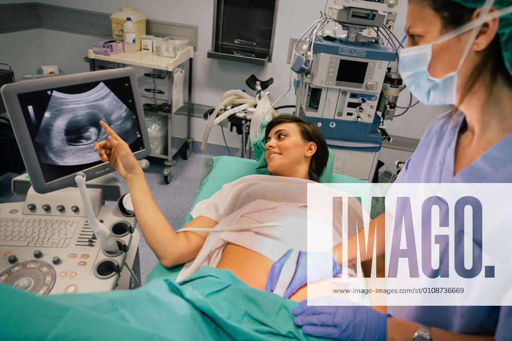 Female doctor in sterile mask and blue glove using ultrasound scanner while examining cheerful pregnant woman pointing at the screen in hospital, Model released, Property released