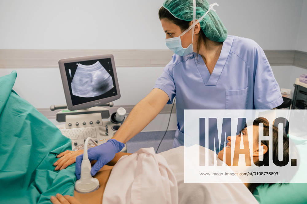 Female doctor in sterile mask and blue glove using ultrasound scanner while examining cheerful pregnant woman in hospital, Model released, Property released