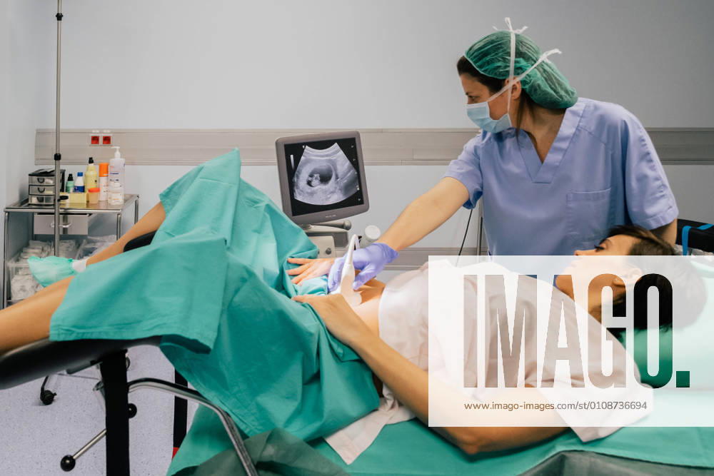 Female doctor in sterile mask and blue glove using ultrasound scanner while examining cheerful pregnant woman in hospital, Model released, Property released