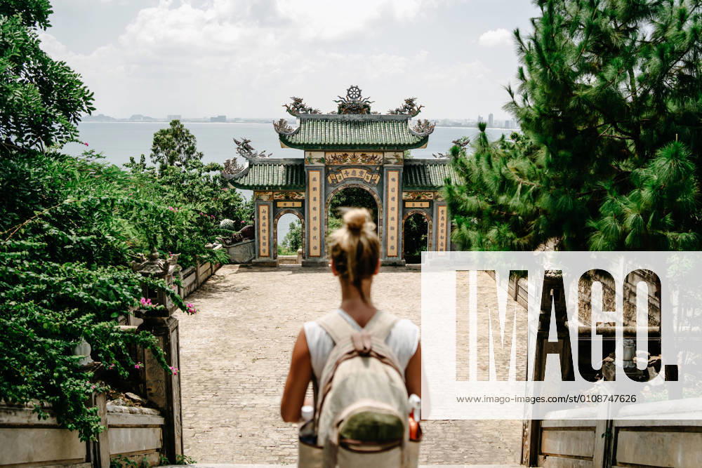 Back view of anonymous female tourist with backpack standing on stairs in front of arch of Buddhist temple in Vietnam