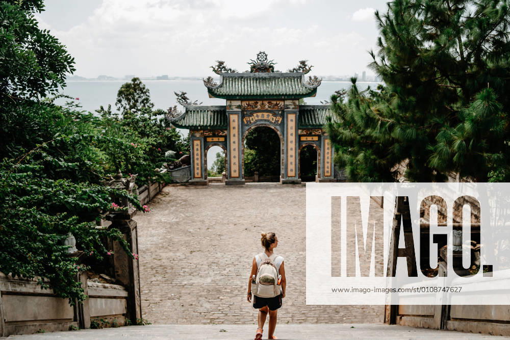 Back view of anonymous female tourist with backpack standing on stairs in front of arch of Buddhist temple in Vietnam
