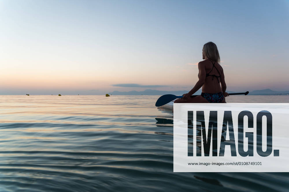 Side view of of unrecognizable female surfer sitting on paddleboard and rowing against spectacular sun in sunset sky