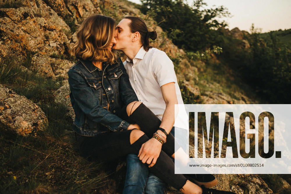 Young man kissing girlfriend in cheek while sitting together on slope of rocky mountain, Model released