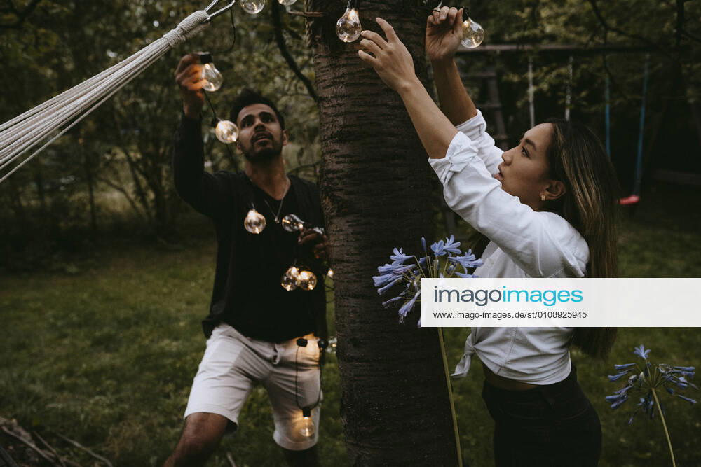 Male and female friends hanging lighting equipment in yard during dinner party Sweden, Stockholm, model released, property released