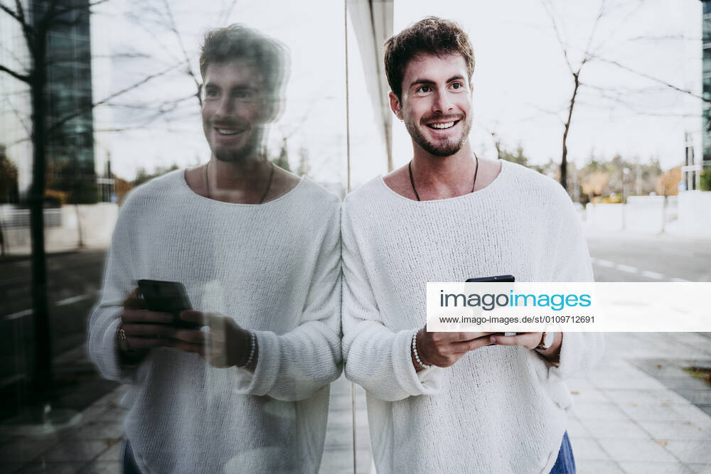 Smiling man with smart phone looking away while standing by building in city model released Symbolfoto property released EBBF01478